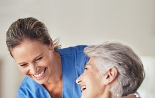 Eine lächelnde Pflegekraft in blauer Uniform umarmt eine ältere Dame, die in einem hellblauen Hemd gekleidet ist. Beide sehen glücklich aus und die ältere Dame hält die Hand der Pflegekraft, während sie in einem hellen, freundlichen Raum sitzt. Die Interaktion vermittelt Wärme, Fürsorge und eine positive Atmosphäre, die den Krankenpflegeförderverein Wadgassen repräsentieren soll
