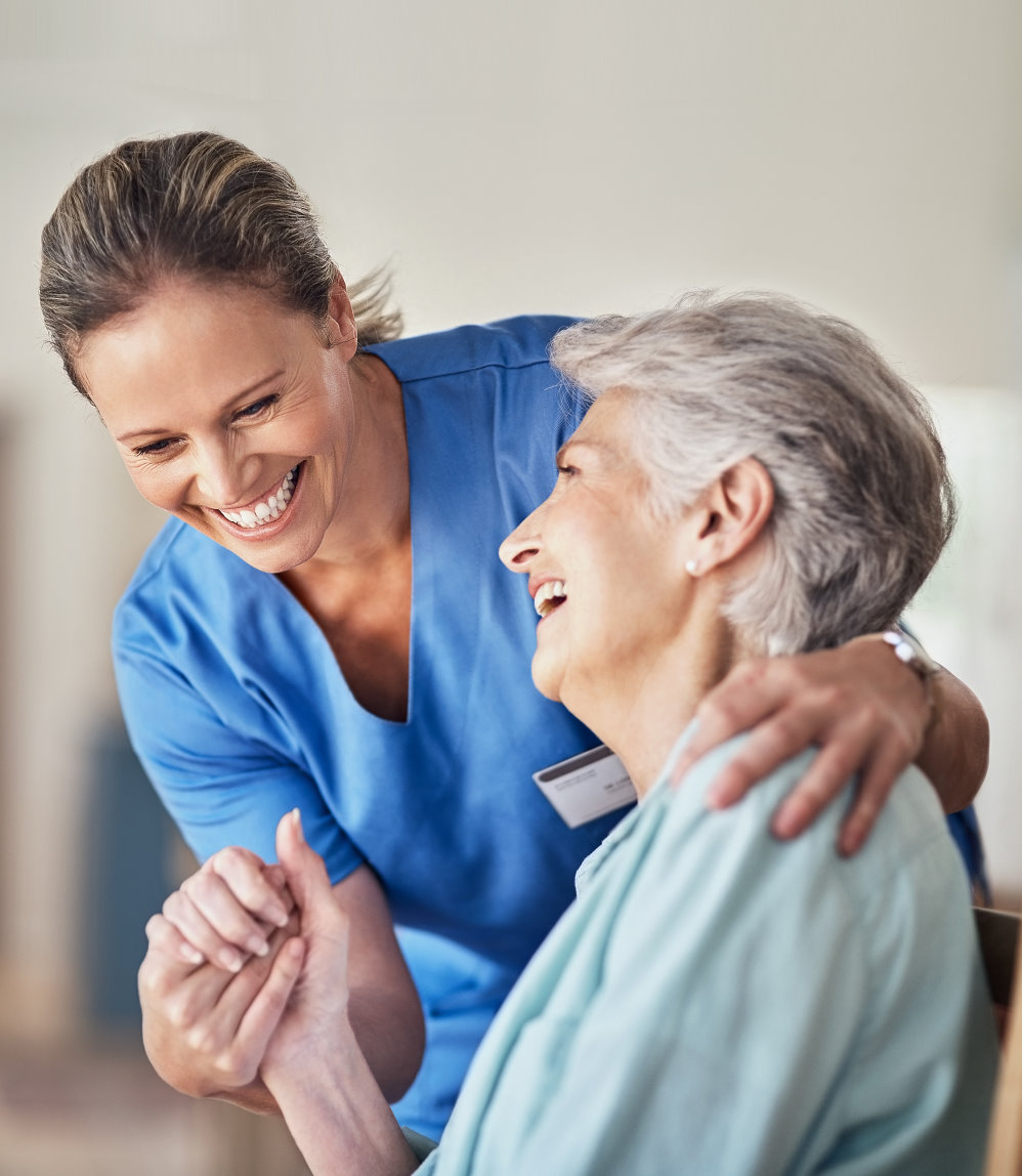 Eine lächelnde Pflegekraft in blauer Uniform umarmt eine ältere Dame, die in einem hellblauen Hemd gekleidet ist. Beide sehen glücklich aus und die ältere Dame hält die Hand der Pflegekraft, während sie in einem hellen, freundlichen Raum sitzt. Die Interaktion vermittelt Wärme, Fürsorge und eine positive Atmosphäre, die den Krankenpflegeförderverein Wadgassen repräsentieren soll